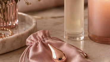 A small smooth rose-gold teardrop jewel ornament resting on a blush silk pouch beside a lubricant bottle and a candle