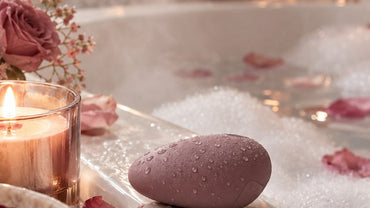 A smooth pebble-shaped mauve silicone wellness device beaded with water droplets on the edge of a white bathtub