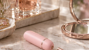 A small smooth lipstick-sized blush pink capsule-shaped wellness device with a rose-gold magnetic charging cable on a marb...