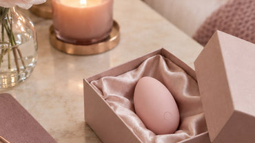 A small smooth egg-shaped blush silicone wellness device nestled in an open satin-lined box on a nightstand with a noteboo...