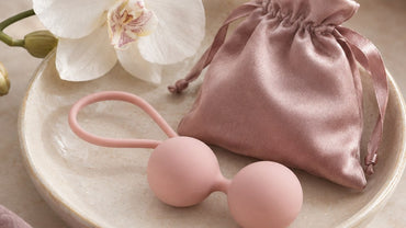 Two small smooth blush silicone spheres connected by a soft loop resting on a pale ceramic dish next to a tiny satin pouch...