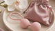 Two small smooth blush silicone spheres connected by a soft loop resting on a pale ceramic dish next to a tiny satin pouch...