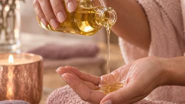 Hands pouring warm golden massage oil from a glass bottle into a palm over a blush towel
