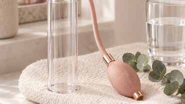 A sleek unbranded clear acrylic cylinder case and a small rose-gold hand pump bulb laid neatly on a folded cream towel bes...