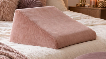 A plush blush velvet wedge cushion and a round mauve upholstered ottoman on a cream linen bed with soft throw pillows