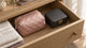 A blush quilted satin zip pouch and a small charcoal lockable case resting in an open oak dresser drawer beside folded linen