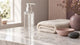 A clear label-free pump bottle of gel on a marble vanity beside a folded cream towel, a blush ceramic dish and a smooth dark stone